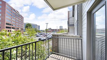 A balcony with a black railing overlooks a parking lot and buildings.